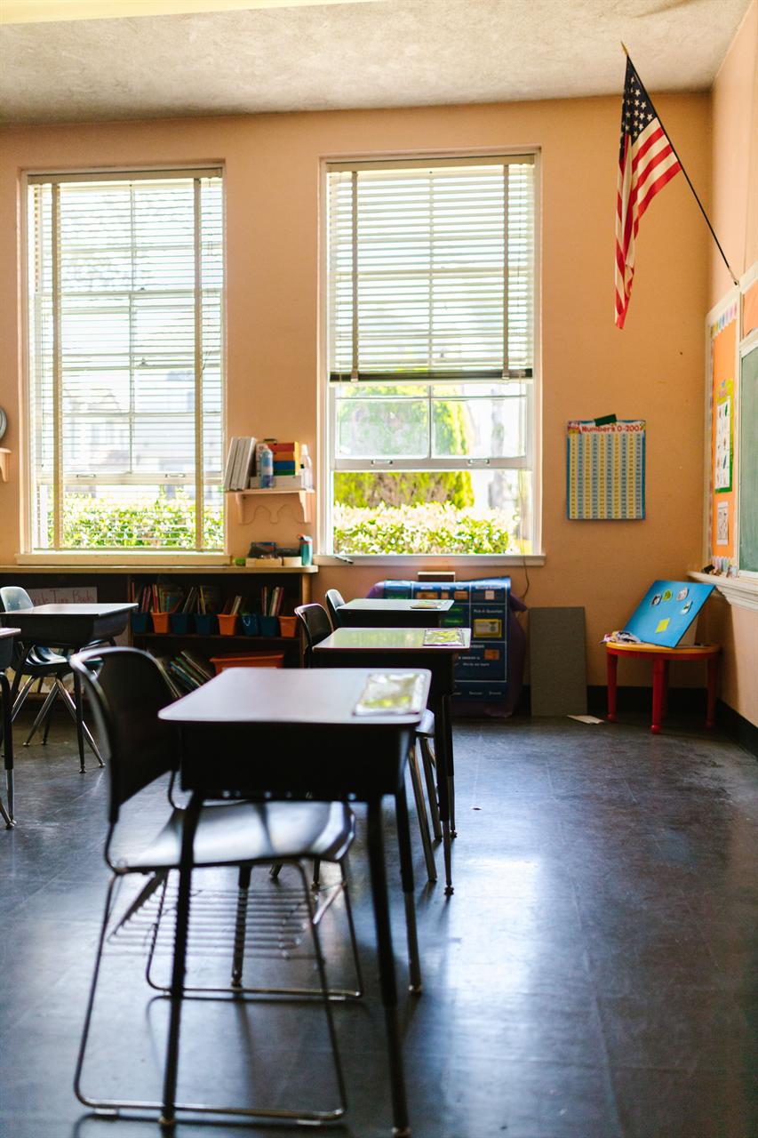 A bright, sunlit K-12 elementary classroom with empty student desks, a bookshelf of supplies, and an American flag